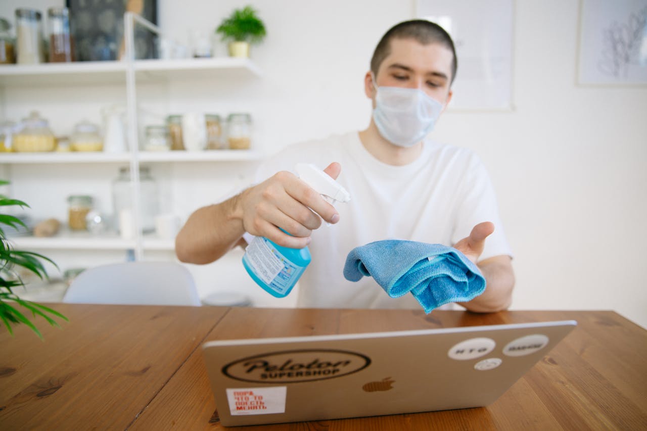 about-02 Man using disinfectant to clean laptop with a face mask on.