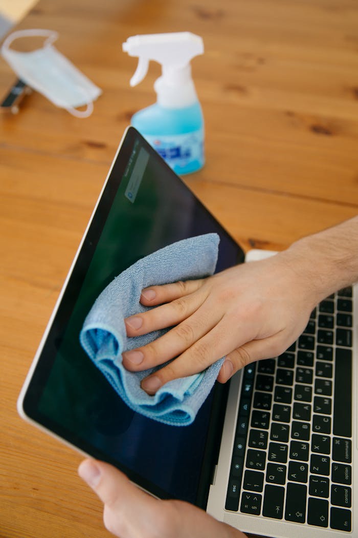 services-01 A person wipes a laptop screen using a blue cloth and spray bottle on a wooden table for hygiene.